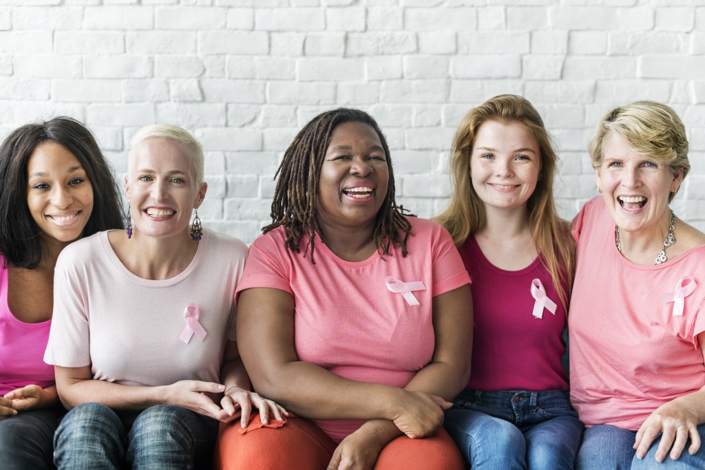 5 women sit in front of a white brick wall smiling. They wear various pink colored shirts and have a small pink ribbon pinned to their shirt. Tags: Breast Cancer Awareness, 3D Mammo, Mammography, Call 844.414.3627 to book your appointment today.